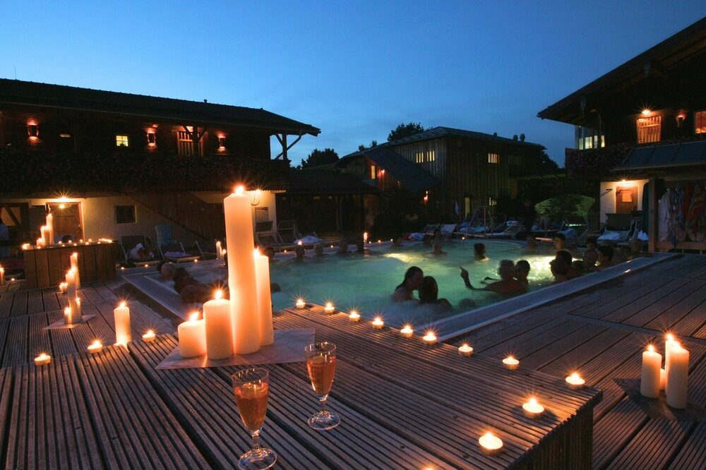 Ein hölzernes Deck mit Kerzen vor einem beleuchteten Pool bei Nacht für ein einzigartiges Saunaerlebniss in Bad Füssing.