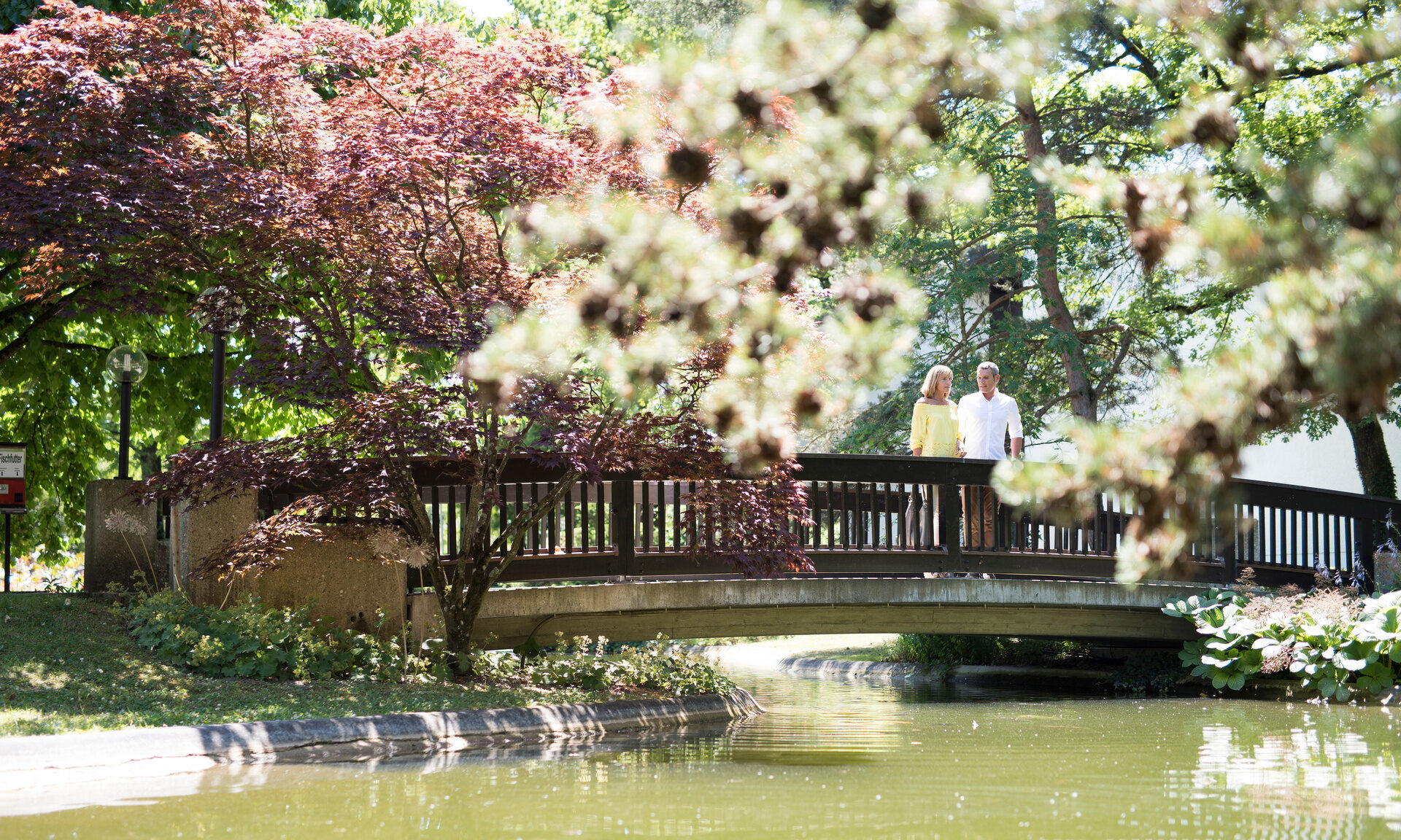 Eine Brücke über einem Wasserlauf im Kurpark Bad Füssing mit blühenden Bäumen im Hintergrund.