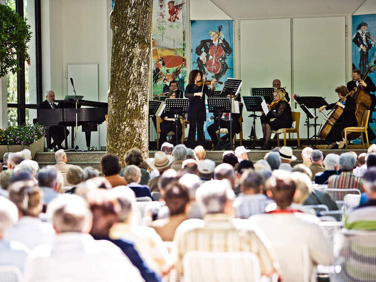 Eine Gruppe von Personen sitzt auf Stühlen in einem Park von Bad Füssing, umgeben von klassischer Musik eines Konzertes.