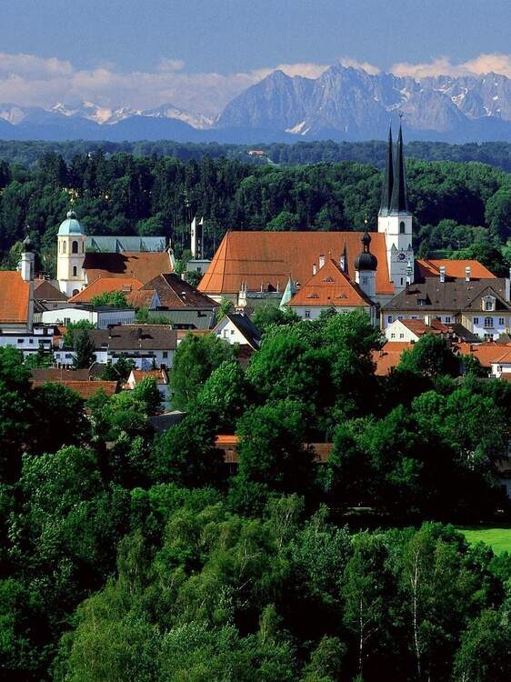 Stadt Altötting mit Kirche nd mehreren Häusern, umgeben von Wäldern, dahinter Gebirge,