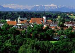 Stadt Altötting mit Kirche nd mehreren Häusern, umgeben von Wäldern, dahinter Gebirge,